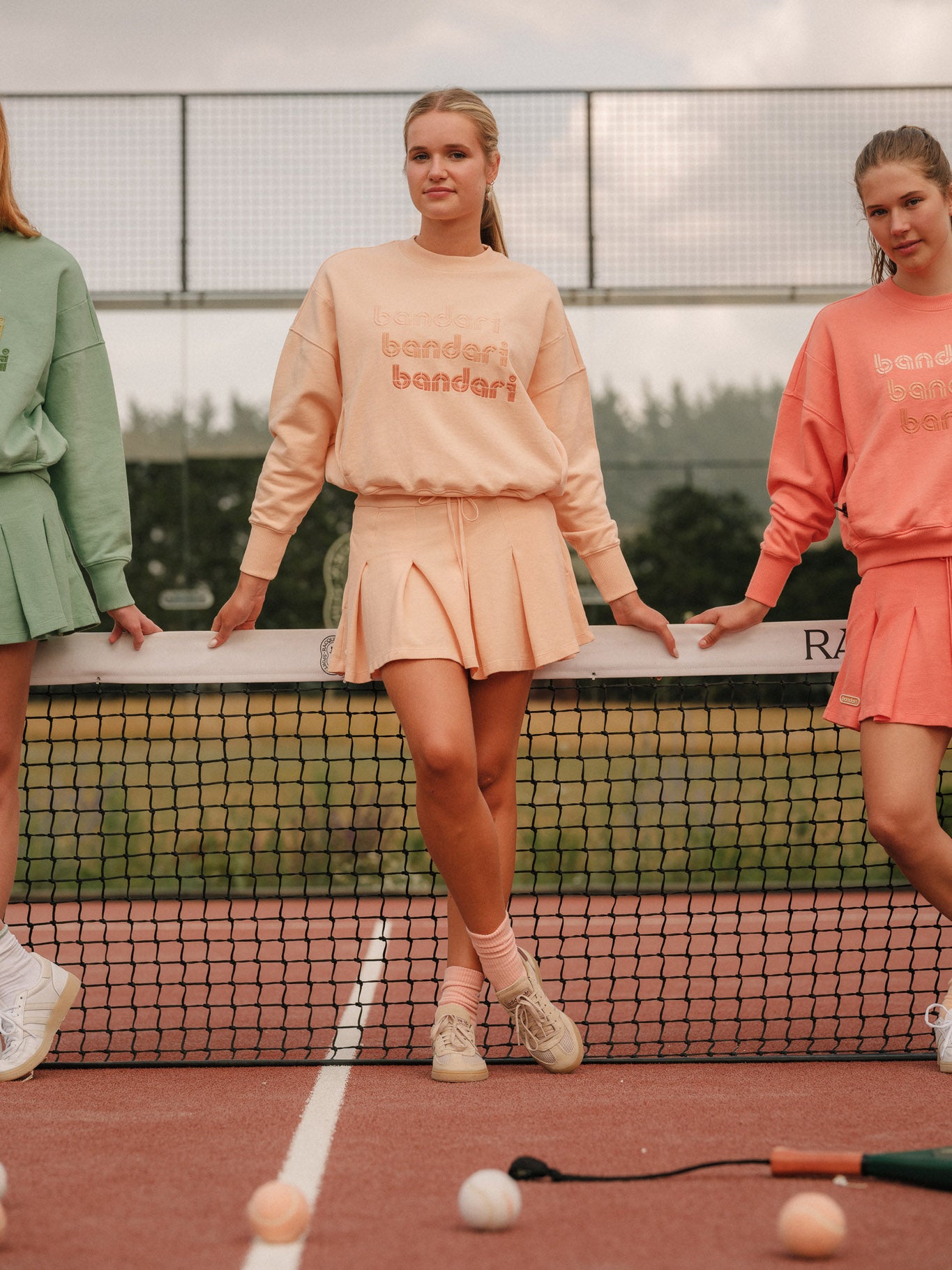 Three models wearing Bandari padel sweatshirts in Faded Peach, Verdant Green and Sunset Orange - women's organic cotton loopback jersey, 3 tone embroidery, front view, worn on padel court with matching coloured skorts